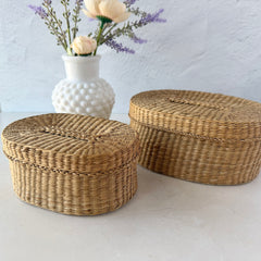 Two oval sweetgrass baskets with lids, hand‑woven in natural tones, shown nested together beside a white vase with flowers.