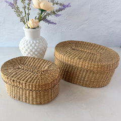 Two oval sweetgrass baskets with lids, hand‑woven in natural tones, shown nested together beside a white vase with flowers.