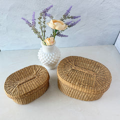 Two oval sweetgrass baskets with lids, hand‑woven in natural tones, shown nested together beside a white vase with flowers.