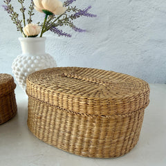 Two oval sweetgrass baskets with lids, hand‑woven in natural tones, shown nested together beside a white vase with flowers.