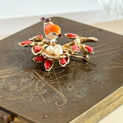 Vintage Swoboda bird nest brooch with a painted bone bird, faux pearl eggs, coral‑colored stone chips, and gold‑tone metal branch detailing.