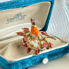 Vintage Swoboda bird nest brooch with a painted bone bird, faux pearl eggs, coral‑colored stone chips, and gold‑tone metal branch detailing.