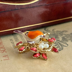 Vintage Swoboda bird nest brooch with a painted bone bird, faux pearl eggs, coral‑colored stone chips, and gold‑tone metal branch detailing.
