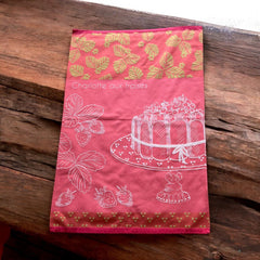 Pink French tea towel with golden strawberry leaves and a white illustration of a Charlotte aux Fraises dessert on a cake stand, by Coucke.