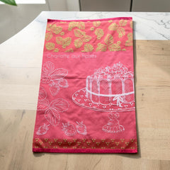 Pink French tea towel with golden strawberry leaves and a white illustration of a Charlotte aux Fraises dessert on a cake stand, by Coucke.