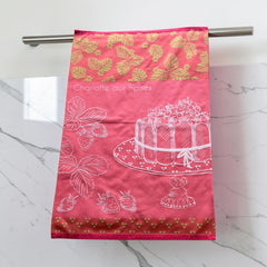 Pink French tea towel with golden strawberry leaves and a white illustration of a Charlotte aux Fraises dessert on a cake stand, by Coucke.