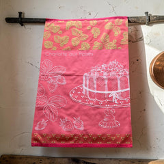 Pink French tea towel with golden strawberry leaves and a white illustration of a Charlotte aux Fraises dessert on a cake stand, by Coucke.