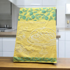 Yellow French tea towel with green lemon and leaf motifs and a white illustration of a lemon meringue tart on a cake stand, by Coucke.