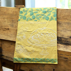 Yellow French tea towel with green lemon and leaf motifs and a white illustration of a lemon meringue tart on a cake stand, by Coucke.