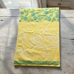 Yellow French tea towel with green lemon and leaf motifs and a white illustration of a lemon meringue tart on a cake stand, by Coucke.