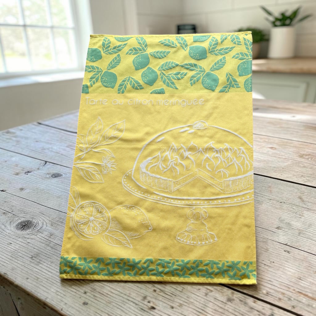 Yellow French tea towel with green lemon and leaf motifs and a white illustration of a lemon meringue tart on a cake stand, by Coucke.