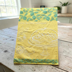Yellow French tea towel with green lemon and leaf motifs and a white illustration of a lemon meringue tart on a cake stand, by Coucke.