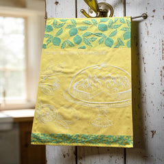 Yellow French tea towel with green lemon and leaf motifs and a white illustration of a lemon meringue tart on a cake stand, by Coucke.