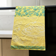 Yellow French tea towel with green lemon and leaf motifs and a white illustration of a lemon meringue tart on a cake stand, by Coucke.