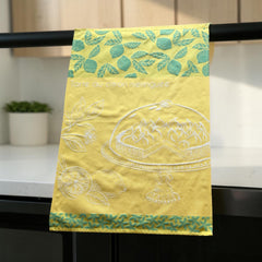 Yellow French tea towel with green lemon and leaf motifs and a white illustration of a lemon meringue tart on a cake stand, by Coucke.