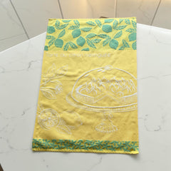 Yellow French tea towel with green lemon and leaf motifs and a white illustration of a lemon meringue tart on a cake stand, by Coucke.