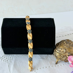 Vintage 1960s Trifari Crown sapphire blue rhinestone birthstone bracelet with brushed gold tone ribbon links and fold‑over clasp.