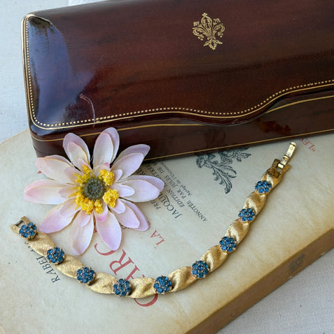 Vintage 1960s Trifari Crown sapphire blue rhinestone birthstone bracelet with brushed gold tone ribbon links and fold‑over clasp.