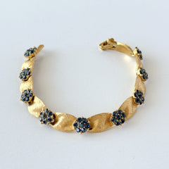 Vintage 1960s Trifari Crown sapphire blue rhinestone birthstone bracelet with brushed gold tone ribbon links and fold‑over clasp.