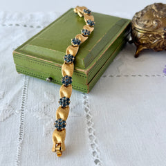 Vintage 1960s Trifari Crown sapphire blue rhinestone birthstone bracelet with brushed gold tone ribbon links and fold‑over clasp.