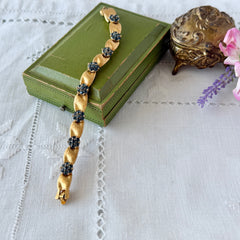 Vintage 1960s Trifari Crown sapphire blue rhinestone birthstone bracelet with brushed gold tone ribbon links and fold‑over clasp.