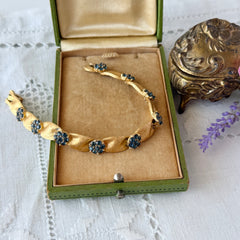 Vintage 1960s Trifari Crown sapphire blue rhinestone birthstone bracelet with brushed gold tone ribbon links and fold‑over clasp.