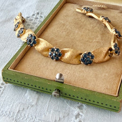 Vintage 1960s Trifari Crown sapphire blue rhinestone birthstone bracelet with brushed gold tone ribbon links and fold‑over clasp.