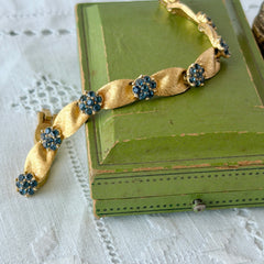 Vintage 1960s Trifari Crown sapphire blue rhinestone birthstone bracelet with brushed gold tone ribbon links and fold‑over clasp.