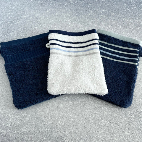Three French cotton terry pocket washcloths in white with blue stripes, navy with blue stripes, and solid navy, arranged on a speckled surface.