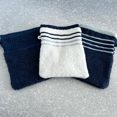 Three French cotton terry pocket washcloths in white with blue stripes, navy with blue stripes, and solid navy, arranged on a speckled surface.