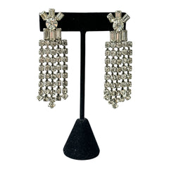 Vintage 1960s clip-on earrings with cascading clear rhinestone dangles, featuring chaton and baguette stones in a waterfall design, displayed on a black stand.