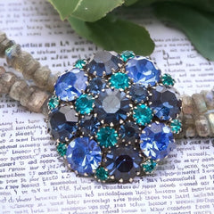  A stunning vintage Weiss crystal dome brooch in shades of blue and teal, paired with a double strand of shiny blue and gray faceted labradorite rondelles. The brooch can be worn centered, off-center, or separately, offering versatile styling. Finished with sterling silver spacers and a toggle clasp, this unique piece blends vintage charm with modern craftsmanship, signed Weiss on the back and featuring the EM tag in Etruscan alphabet for Elisabeth Michel-Meyrueix.
