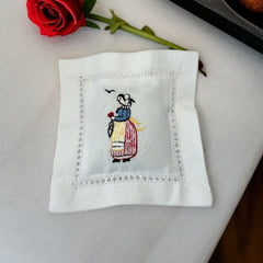 White cotton sachet pillow embroidered with a traditional Breton woman in striped skirt and bonnet, holding a red flower beneath two flying birds. Framed by a lace-style hemstitch border and filled with fragrant French lavender buds, this handmade cushion evokes cultural charm and artisanal heritage.