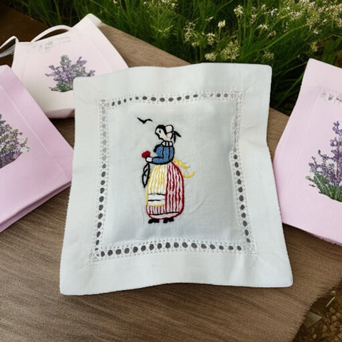 White cotton sachet pillow embroidered with a traditional Breton woman in striped skirt and bonnet, holding a red flower beneath two flying birds. Framed by a lace-style hemstitch border and filled with fragrant French lavender buds, this handmade cushion evokes cultural charm and artisanal heritage.