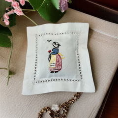 White cotton sachet pillow embroidered with a traditional Breton woman in striped skirt and bonnet, holding a red flower beneath two flying birds. Framed by a lace-style hemstitch border and filled with fragrant French lavender buds, this handmade cushion evokes cultural charm and artisanal heritage.
