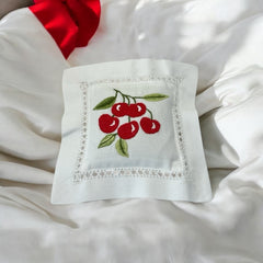 White cotton sachet pillow embroidered with a cluster of red cherries and green leaves, framed by a delicate lace-style hemstitch border. Filled with fragrant French lavender buds, this handmade cushion blends orchard charm with the soothing scent of Provence.