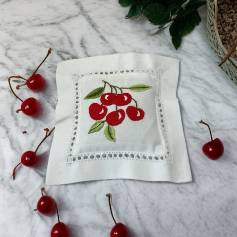 White cotton sachet pillow embroidered with a cluster of red cherries and green leaves, framed by a delicate lace-style hemstitch border. Filled with fragrant French lavender buds, this handmade cushion blends orchard charm with the soothing scent of Provence.