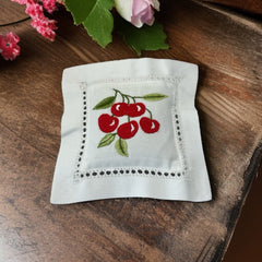 White cotton sachet pillow embroidered with a cluster of red cherries and green leaves, framed by a delicate lace-style hemstitch border. Filled with fragrant French lavender buds, this handmade cushion blends orchard charm with the soothing scent of Provence.