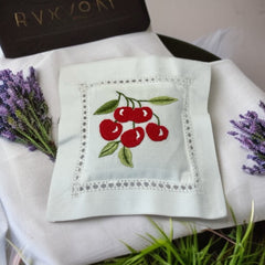 White cotton sachet pillow embroidered with a cluster of red cherries and green leaves, framed by a delicate lace-style hemstitch border. Filled with fragrant French lavender buds, this handmade cushion blends orchard charm with the soothing scent of Provence.