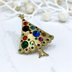 Vintage gold-tone Christmas tree brooch with multicolor rhinestones and scalloped edges, featuring a star at the top and textured detailing; back view shows pin clasp and trunk-shaped base, typical of 1980s–1990s holiday jewelry.